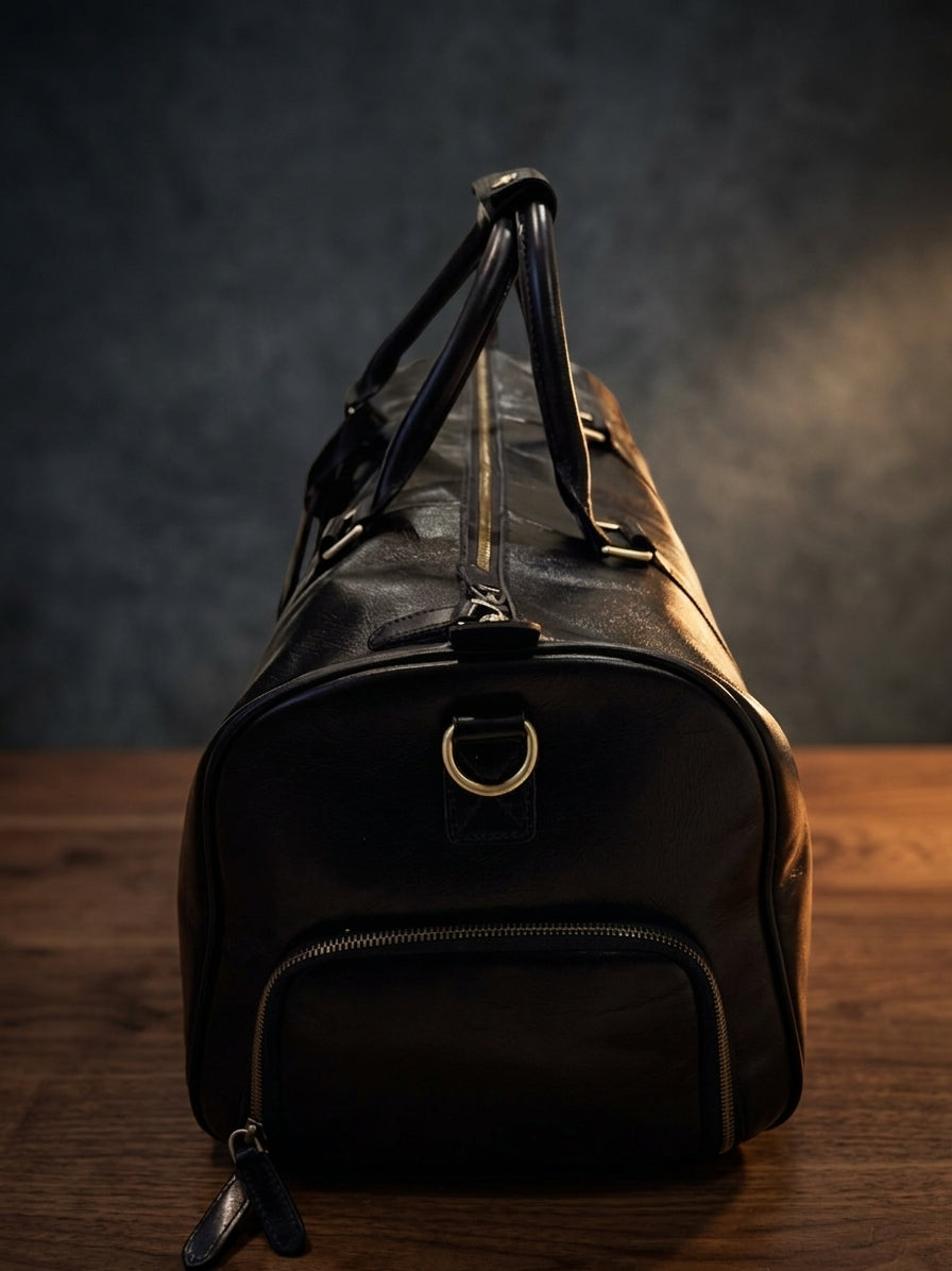 A black leather duffel bag with gold-tone hardware, standing upright on a wooden surface against a dark textured studio background.
